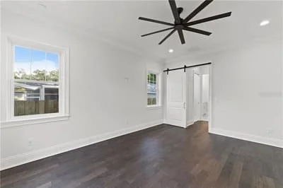 Empty bedroom with dark hardwood floors, white walls, large window, and black ceiling fan